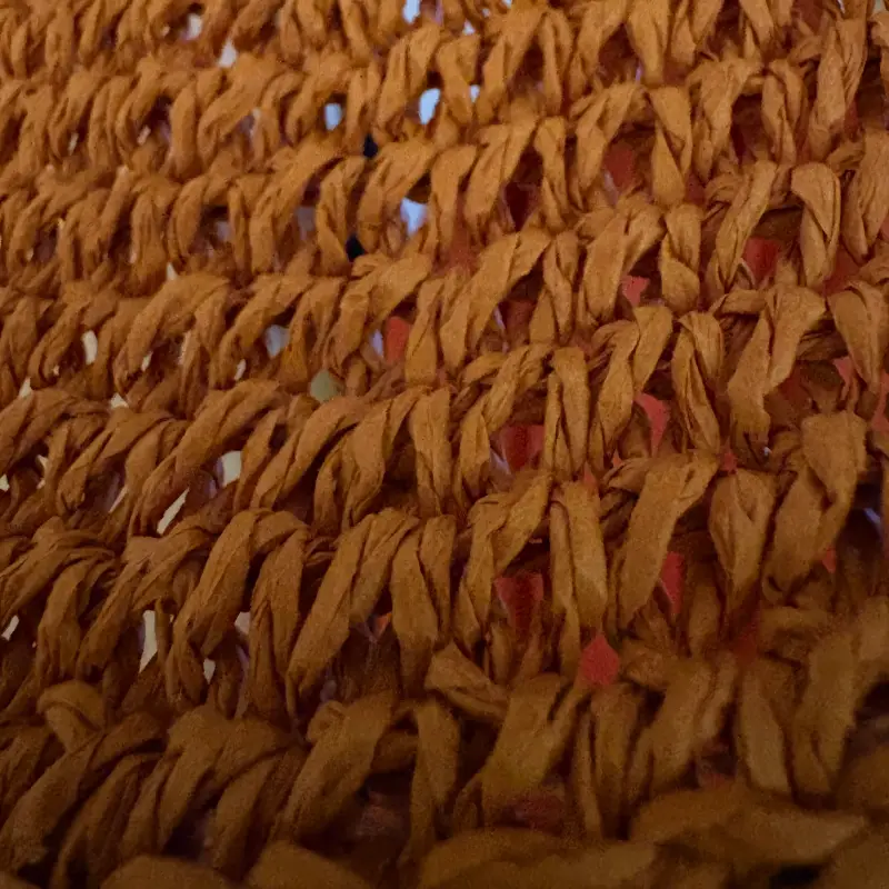 A close-up of open crochet in thick rust-coloured ribbon yarn, its looped rows running across the frame with daylight showing through the gaps