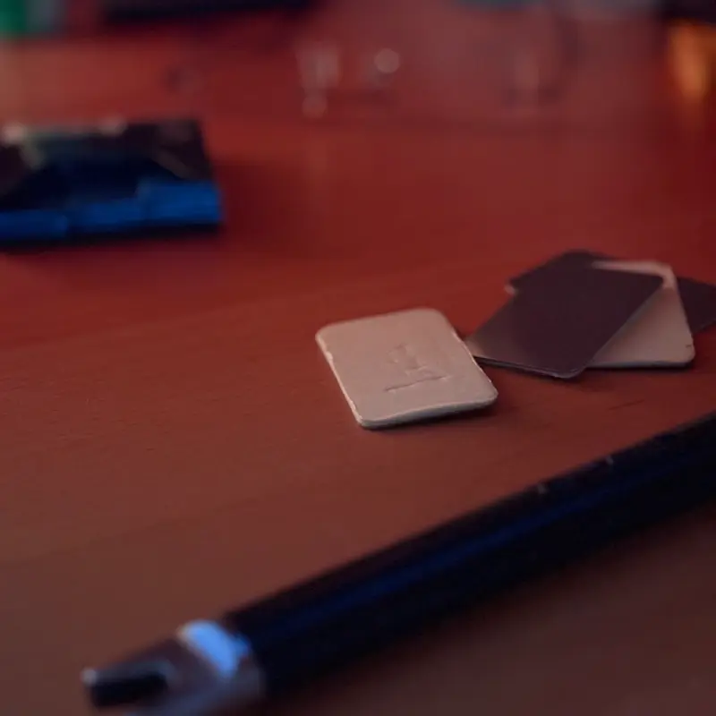 A pale beige tile with an embossed mark on a wooden desk, two dark tiles fanned behind it, a notebook and pen blurred in the foreground