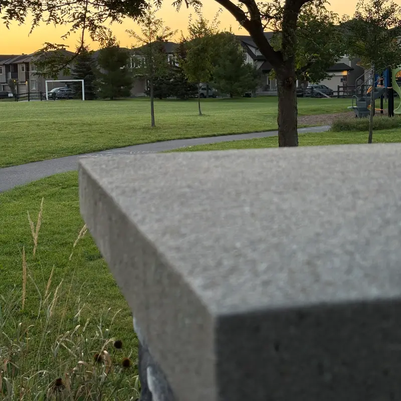 The near corner of a concrete bench slab, far out of focus, cutting diagonally across the lower half of the frame, with a suburban park sharp behind it at dawn: mown grass, an asphalt path curving away, a soccer goal at the left, young staked trees, a playground structure at the right and houses along the skyline under an amber pre-sunrise sky